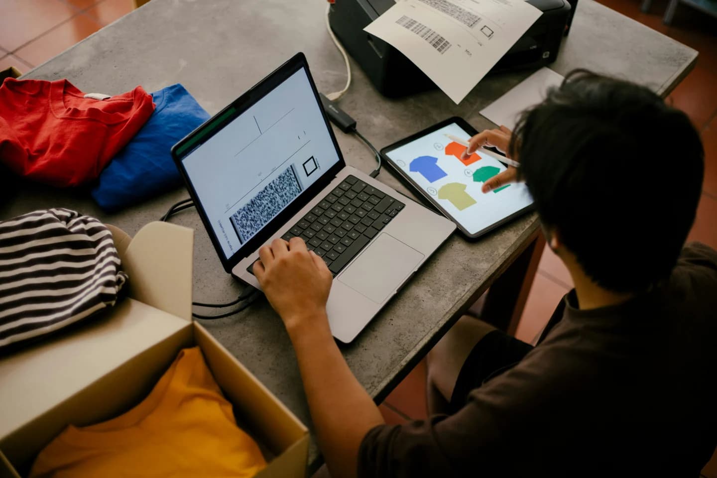 An independent merchant managing orders on a laptop beside clothing and packing materials